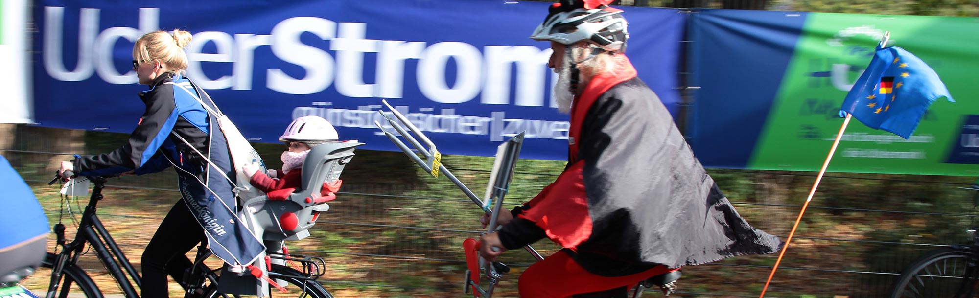 Person in einem rot-schwarzen Kostüm und mit Fahrradhelm, die auf einem Fahrrad an einem blauen Banner vorbeifährt, im Rahmen einer öffentlichen Veranstaltung oder eines Sportevents.
